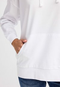 Sudadera blanca con capucha, bolsillo frontal, puños y dobladillo acanalados. Textura de tela suave. Llevada con jeans azules.