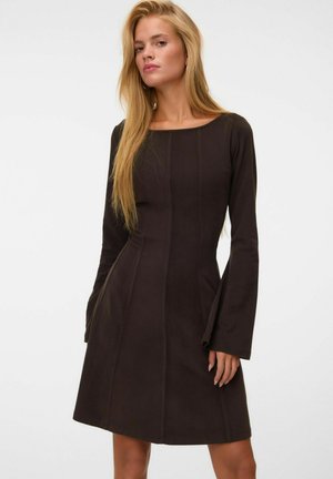 Femme aux longs cheveux blonds portant une robe noire ajustée avec de longues manches évasées sur un fond clair uni.