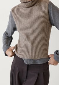 Veste turtleneck tricotée en laine ribbed marron clair, associée à une chemise à manches longues grise en dessous et à un pantalon sombre. Design texturé et superposé.