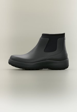Botte de pluie noire montant à la cheville avec une semelle épaisse, des panneaux latéraux élastiques et une finition mate et lisse sur un fond uni.