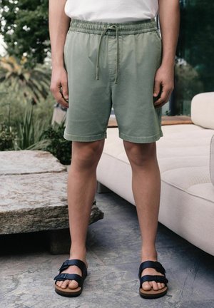 Person wearing light green drawstring shorts and black buckle sandals standing on stone patio near outdoor sofa.