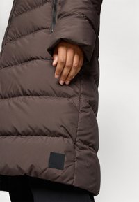 Brun pufferjakke med horisontale sømmer, med glidelås foran og en svart logo-lapp på kanten. Myk, glatt tekstur.