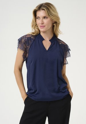Blouse marine avec des manches courtes en dentelle, présentant un col en V et un col froncé, fabriquée à partir d'un tissu lisse, portée avec un pantalon noir.