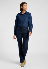 Mörk denimskjorta med knäppning, långa ärmar; kombinerat med mörkblå jeans med lätt flare vid fållen, och beige ankelstövlar.