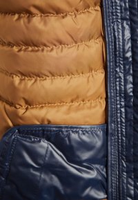 Kurtka pikowana z wnętrzem w kolorze złoto-brązowym oraz zewnętrzną warstwą w teksturze ciemnoniebieskiej, wyposażona w boczną kieszeń o gładkim wykończeniu.