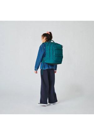 Junge Person mit einem türkisfarbenen Rucksack und einer Jeansjacke, steht und schaut seitlich vor einem einfarbigen hellen Hintergrund.