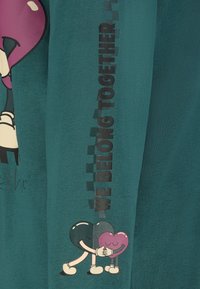 Türkisfarbenes Langarmshirt mit cartoonhaften Herzen, die sich umarmen, und dem Text "WIR GEHÖREN ZUSAMMEN", der vertikal in Schwarz entlang des Ärmels aufgedruckt ist.