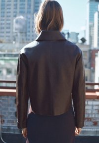 Brązowa skórzana krótka kurtka z szerokim kołnierzem, gładką fakturą i czystymi liniami, widoczna od tyłu na tle miejskim.