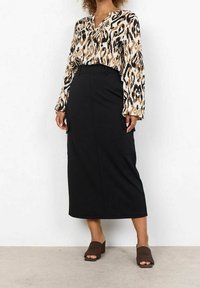 Longue jupe noire avec deux poches latérales, accompagnée d'un blouse beige et noire à motif, dotée de manches amples et d'un décolleté en V.