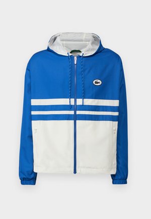 Chaqueta con capucha azul y blanca con cremallera, que presenta dos rayas blancas en el pecho, puños elásticos y un pequeño logo en el lado izquierdo.