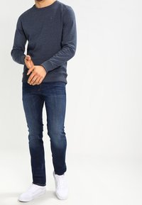 Mörkblå tröja med rund hals, långa ärmar och texturerat tyg. Matchad med mörka jeans och vita sneakers, står med händerna framme.