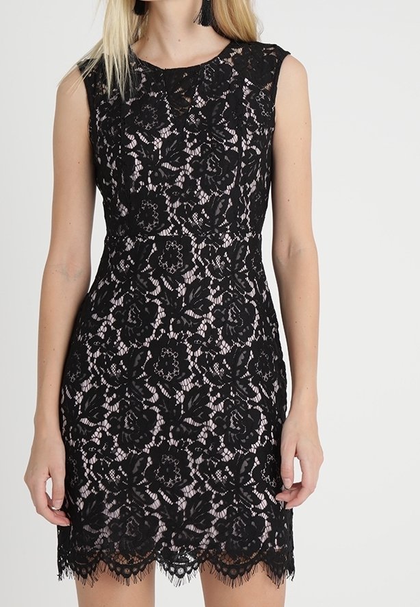 Femme portant une robe noire en dentelle sans manches avec motif floral sur une doublure rose clair, debout devant un fond uni.
