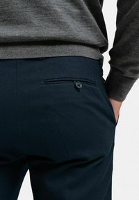 Mörkblå byxor i textil med struktur, som har en bakficka med knapp och en accenttråd. Matchas med en grå tröja.