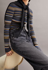 Pull en tricot à rayures multicolores avec des tons de bleu, vert et marron ; jeans gris avec une finition texturée ; pull drapé autour du cou.