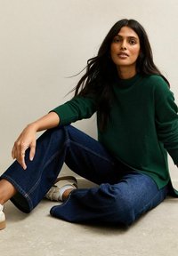 Zielony sweter robiony na drutach z okrągłym dekoltem i ściągaczami na rękawach. Luźno dopasowane ciemnoniebieskie dżinsy. Białe trampki z beżowymi podeszwami.