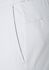 Ljust grått tyg med subtil textur, som har en sidoficka och prydlig sömnad. Designen är slät och figurnära.