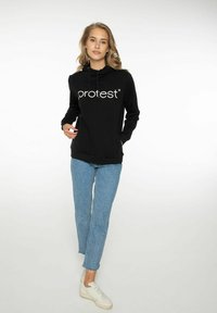 Sudadera negra con cordón y gran logo blanco "pro:test", combinada con vaqueros de mezclilla azul claro y zapatillas blancas. Textura de tela suave.