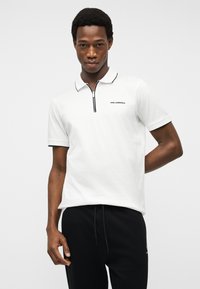 Jeune homme portant un polo blanc à fermeture éclair avec des bordures noires et un pantalon noir, debout devant un fond clair uni.