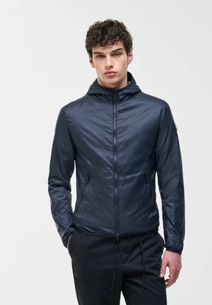 Mladý muž s kudrnatými vlasy, oblečený v tmavě modré zipové mikině s kapucí a černých kalhotách, stojící s jednou rukou v kapse na bílém pozadí.