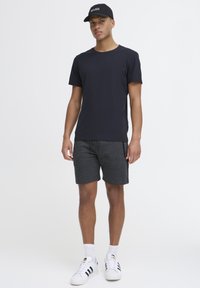 Dunkelblaues T-Shirt mit kurzen Ärmeln, kombiniert mit grauen Shorts. Weiße Sportschuhe mit schwarzen Streifen und eine schwarze Cap mit Aufschrift.