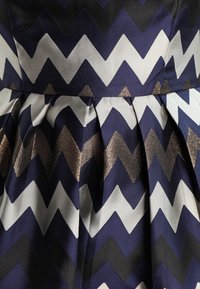 Marinblått tyg med zigzag-mönster i nyanser av marinblått, vitt och metalliskt brons; plisserad design med slät textur.