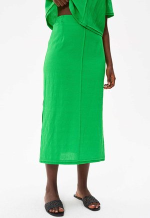 Jupe midi en lin vert à coupe droite, avec des fentes latérales pour faciliter le mouvement et une texture lisse, associée à des sandales tressées noires.
