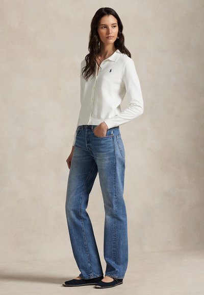 Bela jopica s gumbi, modre kavbojke z ravnimi hlačnicami, črne balerinke z belo obrobo in majhen logotip na jopici. Gladke teksture, sproščen kroj.