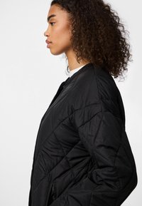 Giacca bomber nera trapuntata con finitura testurizzata, con scollatura tonda e cuciture oblique. Mostrata su un modello in vista laterale.