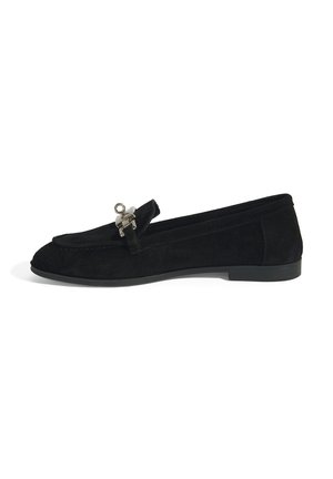 Zwarte suède loafer met een ronde neus, voorzien van een zilverkleurig hardwareaccent aan de voorkant. Platte rubberen zool, strak ontwerp, minimalistische stijl.