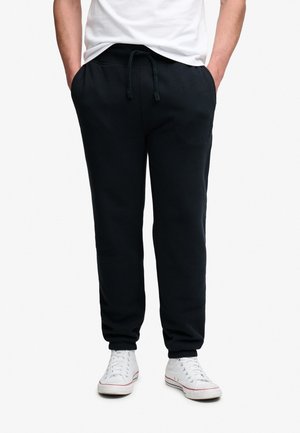 Mand iført sorte joggingbukser med snøre i taljen og hvide høje sneakers, hænderne i lommerne, stående mod en hvid baggrund.