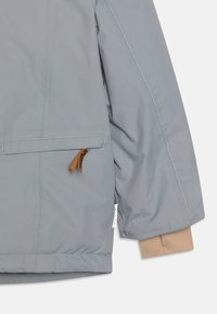 Szara izolowana kurtka z kieszenią na klatce piersiowej, ozdobioną skórzanymi akcentami i beżowym mankietem. Gładka powierzchnia z kontrastującymi detalami ściegów.