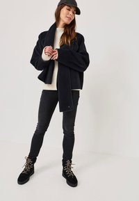 Giacca in pile navy con maniche larghe, indossata sopra un maglione crema. Jeans skinny neri e stivali neri con lacci e dettagli dorati completano il look.