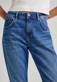 Blå denimjeans med medelhög midja, femficksdesign och subtila blekningar. Har knappstängning och kontrasterande sömmar.