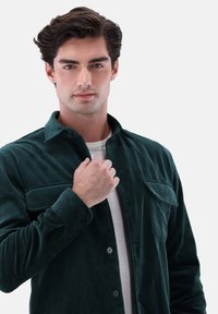 Dark green corduroy shirt with a button-up front, two chest pockets, and a stand collar, featuring a soft texture and ribbed pattern.