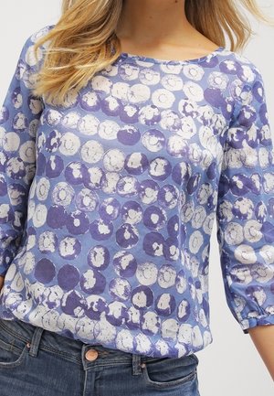 Frau trägt ein blaues Langarmshirt mit weißen und dunkelblauen kreisförmigen Mustern, kombiniert mit dunkelblauen Jeans.