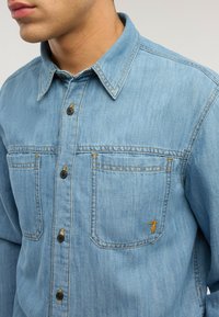 Hombre con camisa de mezclilla azul claro con botones al frente y bolsillos en el pecho con costuras, uno con un pequeño logo bordado amarillo.