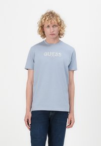 Vaaleansininen puuvillapaita, jossa on pyöreä kaula-aukko ja lyhyet hihat. Paidassa on edessä valkoisella kirjoitettu "GUESS EST. 1981".
