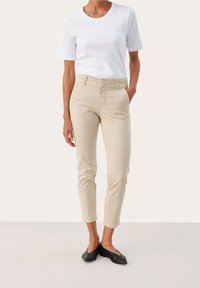 Pantalon en coton beige à coupe ajustée, avec une ceinture, des poches latérales et des poignets retroussés, porté avec un t-shirt blanc à col rond.