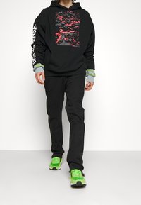 Svart hoodie med röd och grå kamouflagemönster på framsidan, kombinerad med svarta byxor och gröna skor. Har grå och gröna detaljer på ärmsluten.