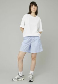 T-shirt blanche à manches courtes, associée à un short rayé bleu clair et des chaussures de sport argentées, se tenant devant un fond neutre.