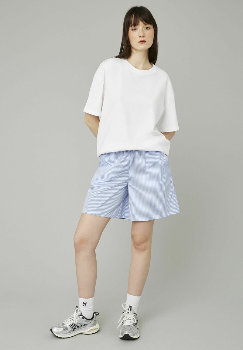 T-shirt blanche à manches courtes, associée à un short rayé bleu clair et des chaussures de sport argentées, se tenant devant un fond neutre.