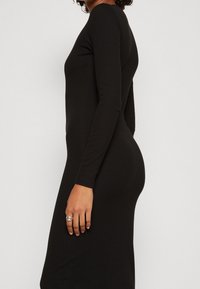 Långärmad svart klänning med en figurnära silhuett, gjord av mjukt material, med rund halsringning och slät yta. Silverring på handen.