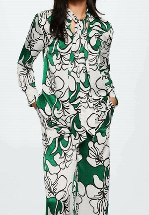 Femme portant un chemisier à motifs floraux verts et blancs avec un col à nouer et un pantalon large assorti, mains dans les poches.