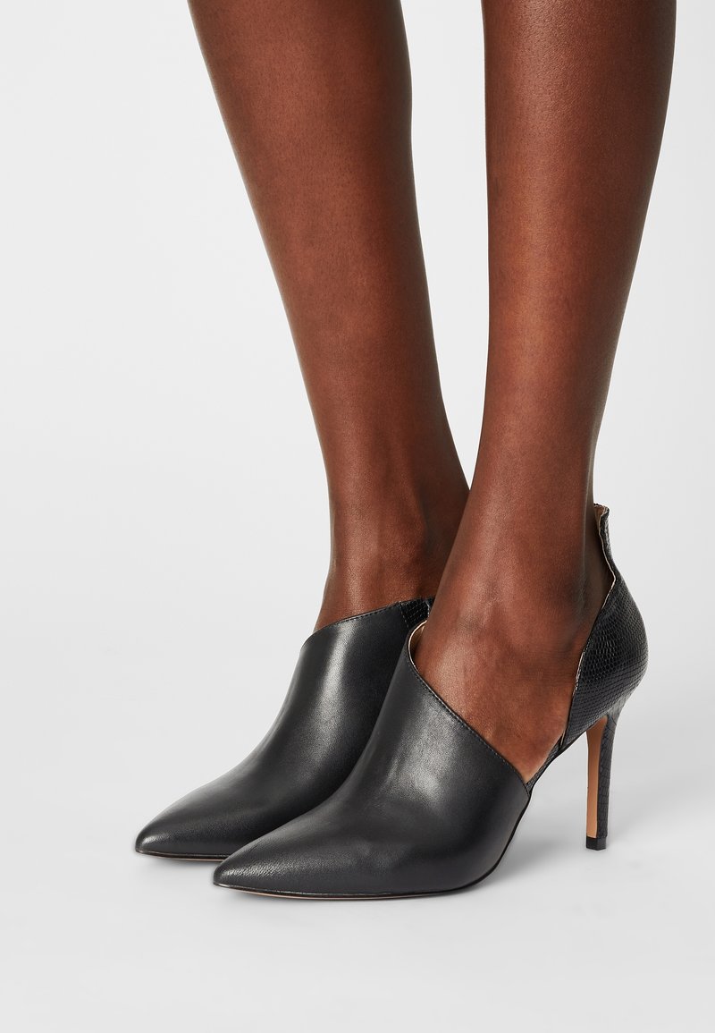 Pieds portant des chaussures à talons aiguilles noirs à bout pointu avec un devant lisse et un panneau arrière texturé sur un fond uni.