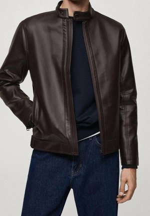 Mężczyzna ubrany w ciemnobrązową skórzaną kurtkę na granatowym swetrze i niebieskich dżinsach, z jedną ręką w kieszeni kurtki, drugą u boku.