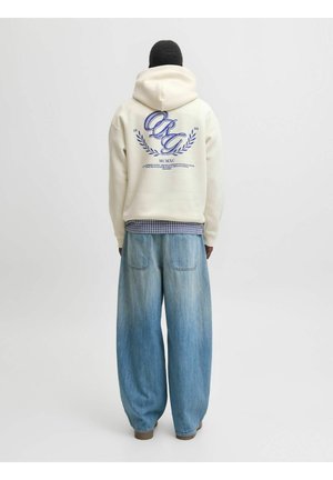 Personne portant un sweat à capuche crème avec un texte graphique bleu dans le dos et un jean bleu clair ample, debout devant un fond uni.