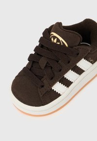 Hnědá semišová teniska s bílým trojpruhovým vzorem, zavazováním vpředu, polstrovaným límcem a oranžovou gumovou podrážkou. Zlaté logo na jazyku.