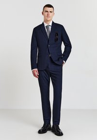 Mann in einem navyblauen karierten Anzug mit passender Hose, weißem Hemd, grauer Krawatte, schwarzen Schuhen und Sonnenbrille, die an der Jackentasche hängt.