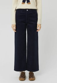 Pantalons larges en velours côtelé bleu marine avec poches avant visibles, fermeture à bouton et nervures texturées ; associés à des chaussures à lacets marron.