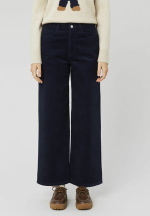 Pantalons larges en velours côtelé bleu marine avec poches avant visibles, fermeture à bouton et nervures texturées ; associés à des chaussures à lacets marron.
